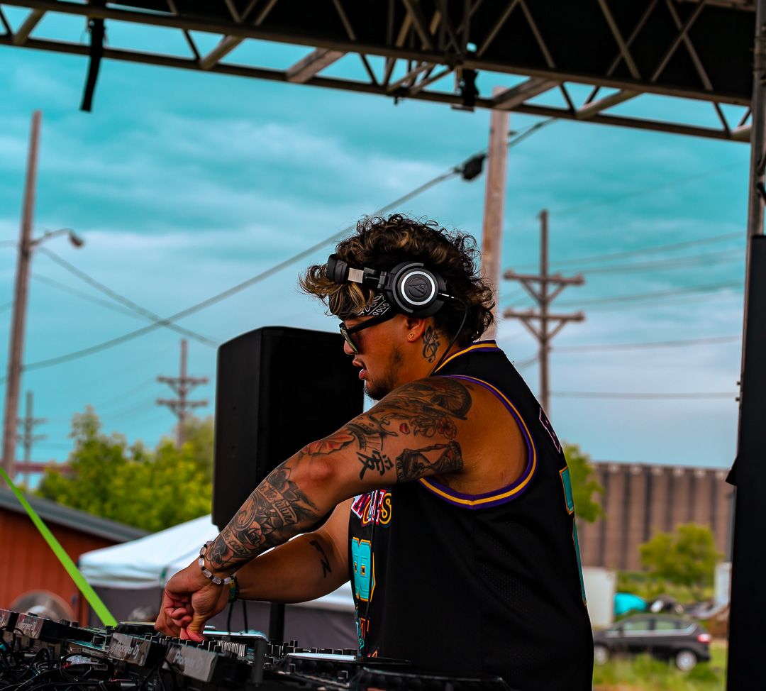 Tattooed DJ performing outdoors at festival, wearing headphones and sunglasses, working turntables under covered stage.