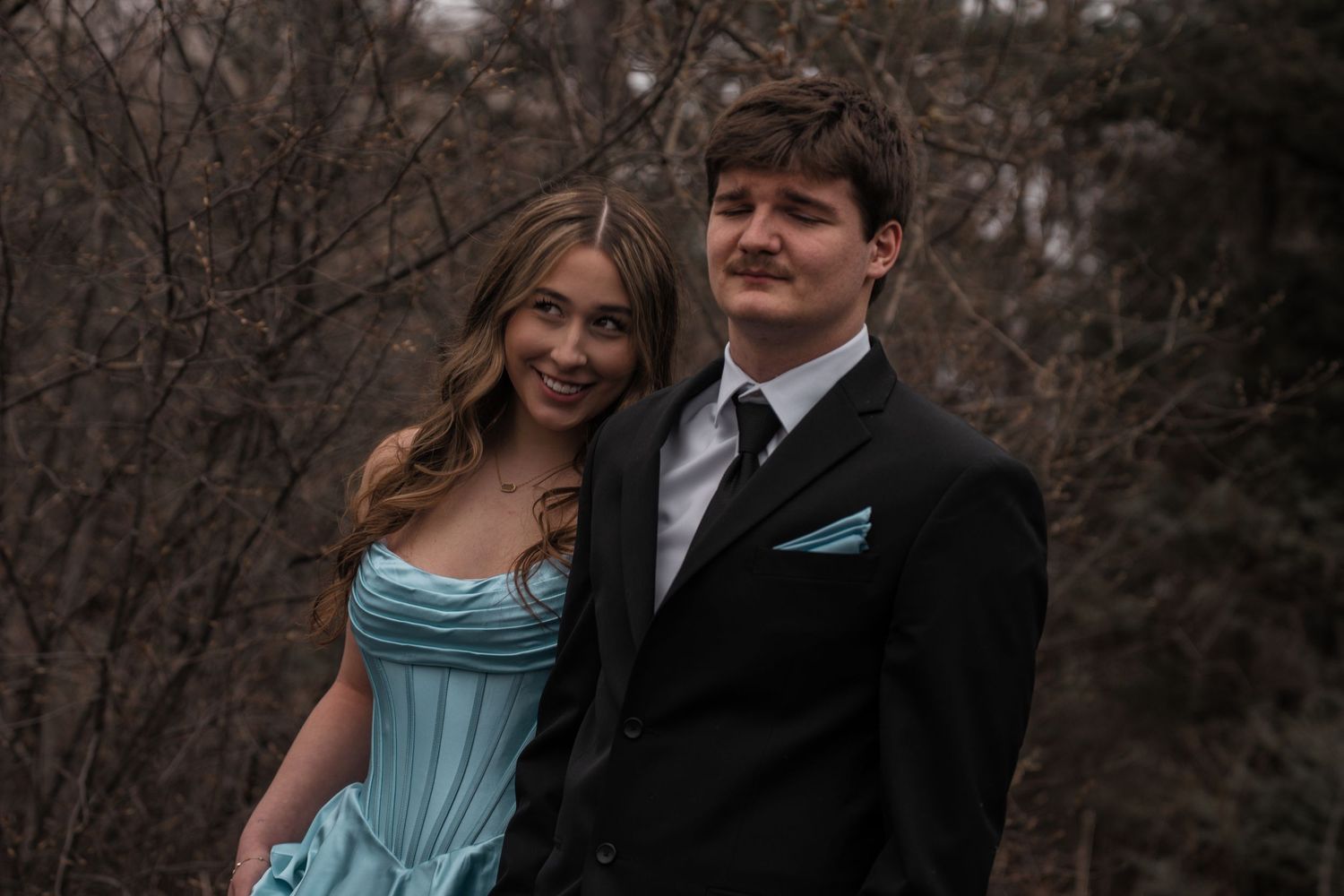 A young couple in formal attire, girl in blue gown and boy in black suit, posing outdoors for prom photos.