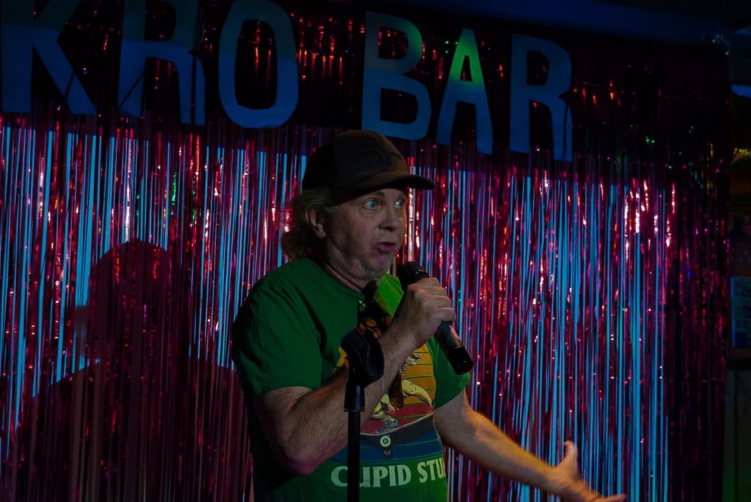 Man in green shirt and hat singing karaoke at KHO BAR with red and blue tinsel curtain backdrop.