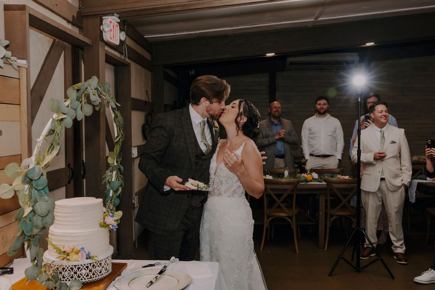 Bride and groom share a kiss while cutting their white wedding cake at a rustic indoor reception venue.
