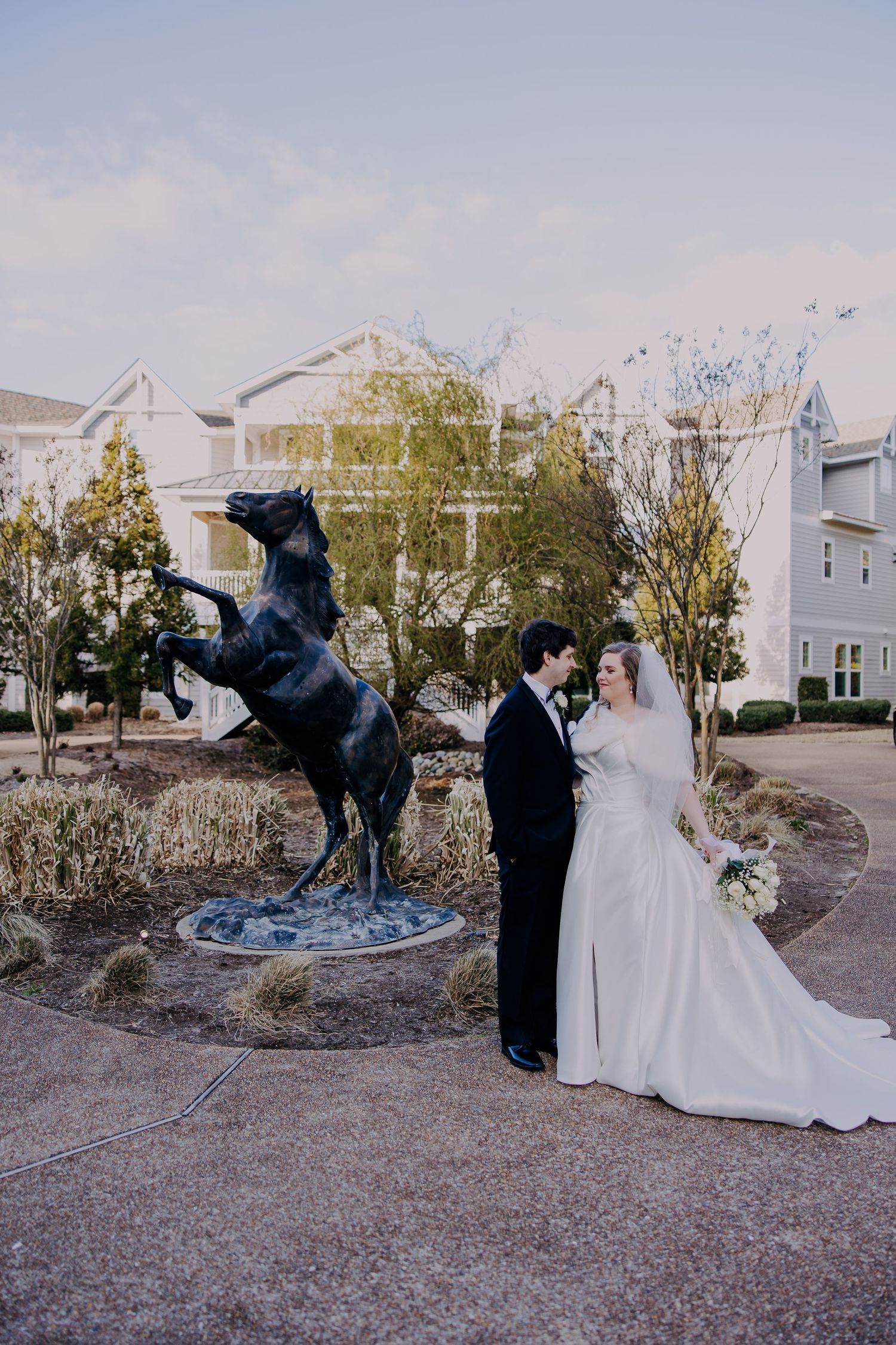 Bride and groom pose beside a rearing horse bronze statue outside a wedding venue at dusk.