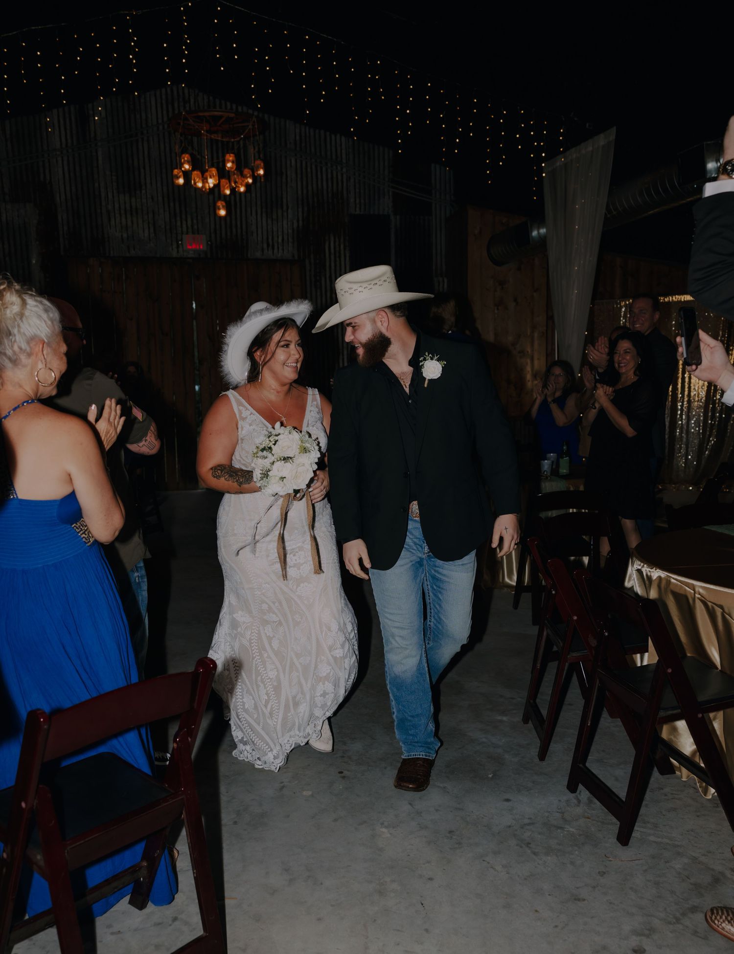 Bride and groom in cowboy hats walk down the aisle at a rustic barn wedding reception with string lights.