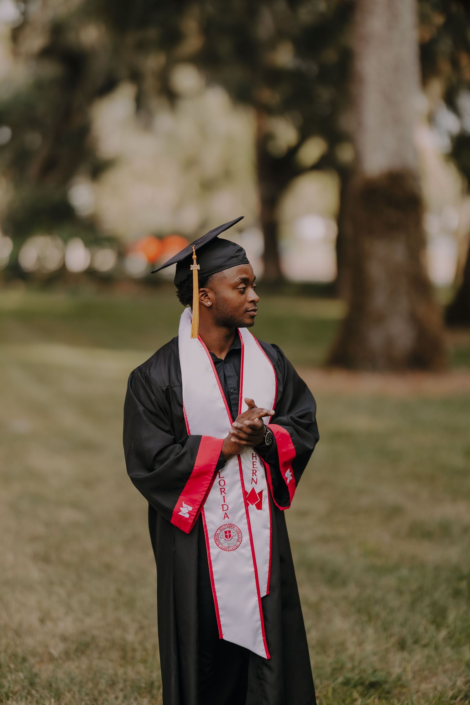 A young man in black graduation cap and gown with red and white stole stands outdoors among trees.