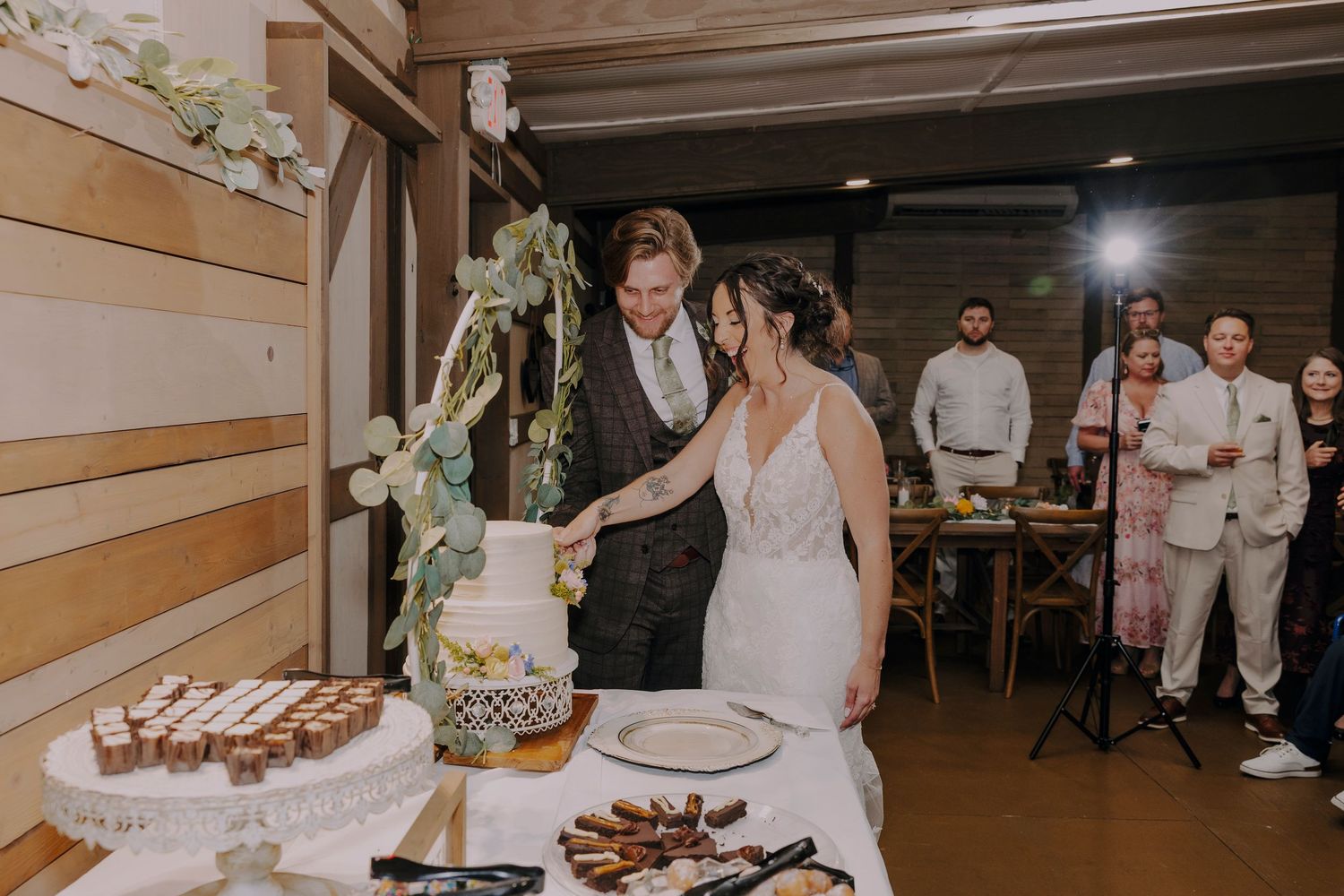 Bride and groom cutting wedding cake together at rustic venue reception with guests watching in background.