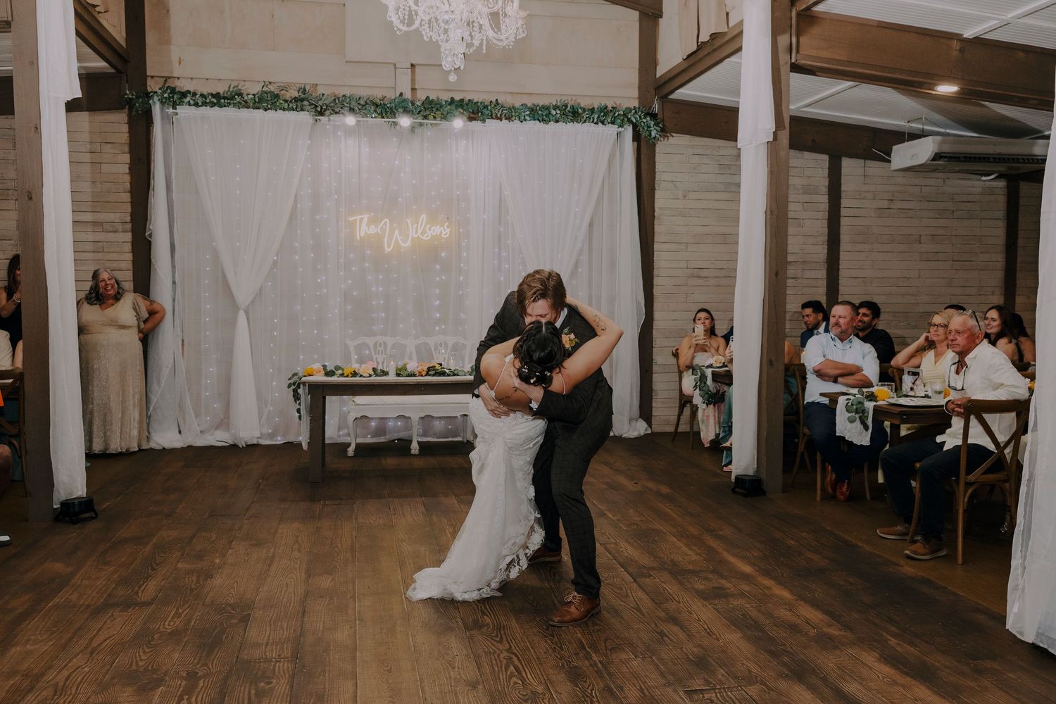 Bride and groom share their first dance in a rustic wedding venue with wood floors and elegant white draping.