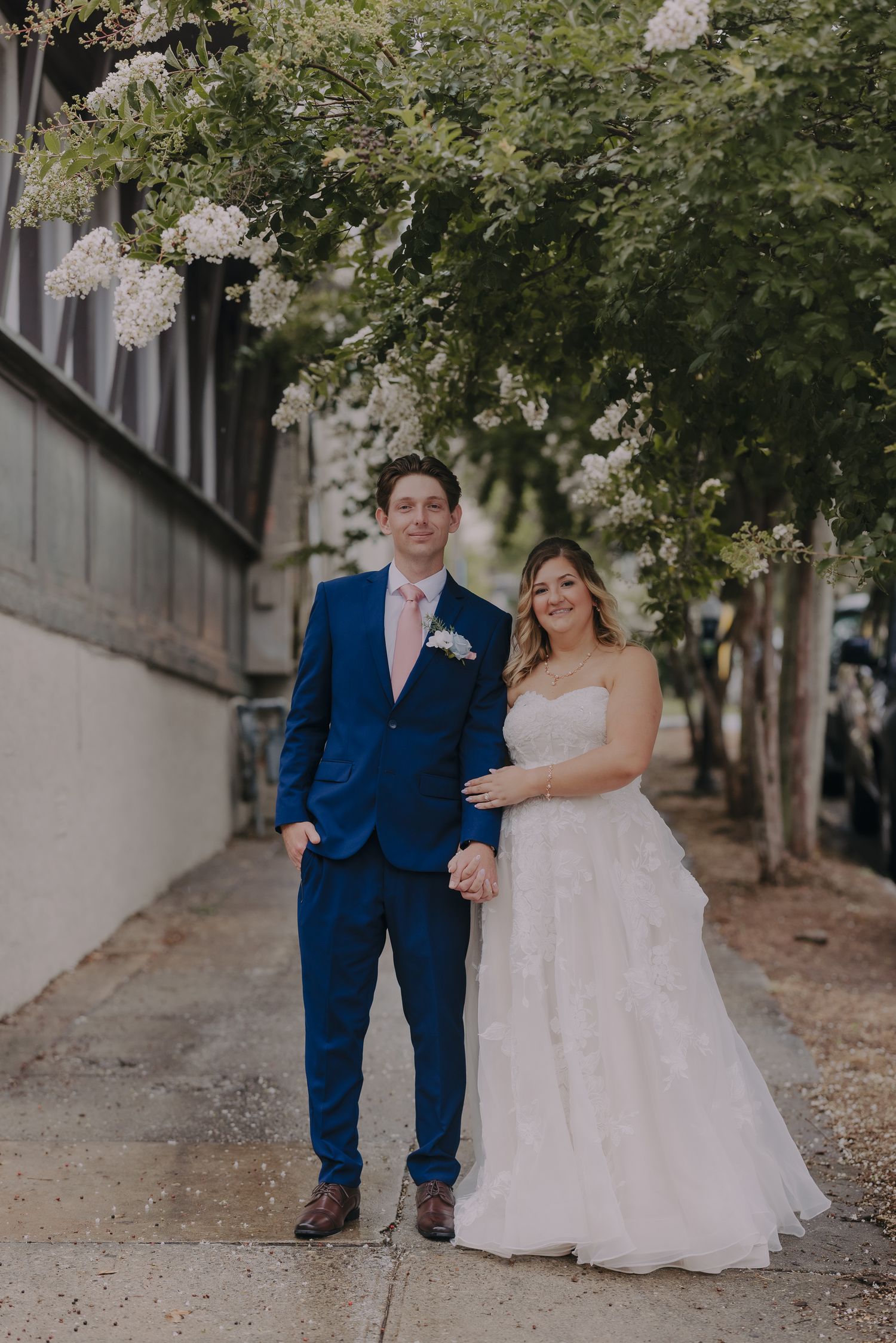 Bride in white strapless gown and groom in blue suit with pink tie pose on sidewalk under blooming white trees.