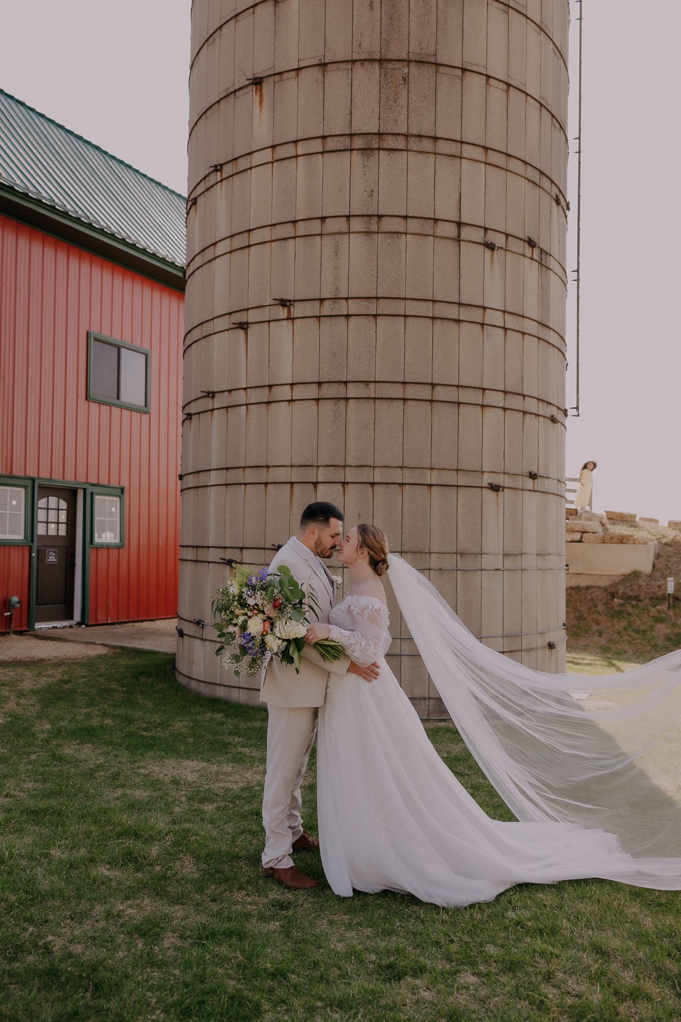 Bride and groom share a romantic kiss in front of a rustic silo at a barn wedding venue, veil flowing in the breeze.