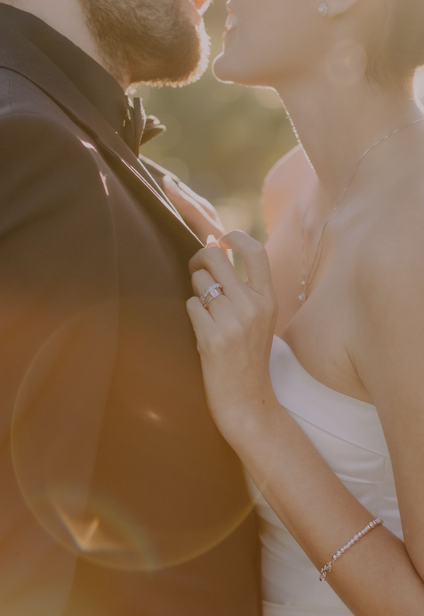 A bride in a white gown with a diamond ring and bracelet gently holds her groom's lapel during a golden hour wedding photo.