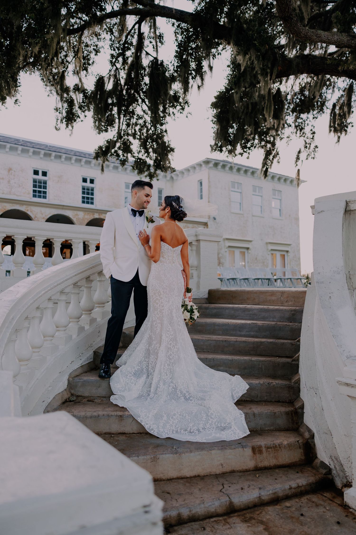 Bride and groom share a romantic moment on elegant white stone stairs outside a grand estate venue.