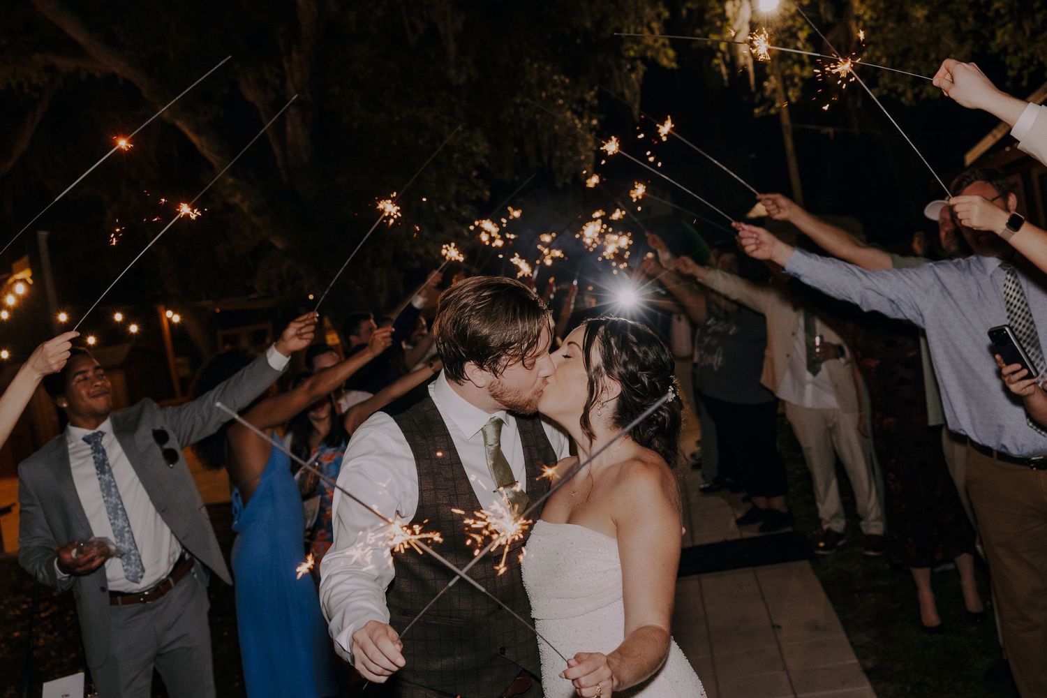 Bride and groom share a kiss during a romantic sparkler send-off surrounded by cheering wedding guests at night.