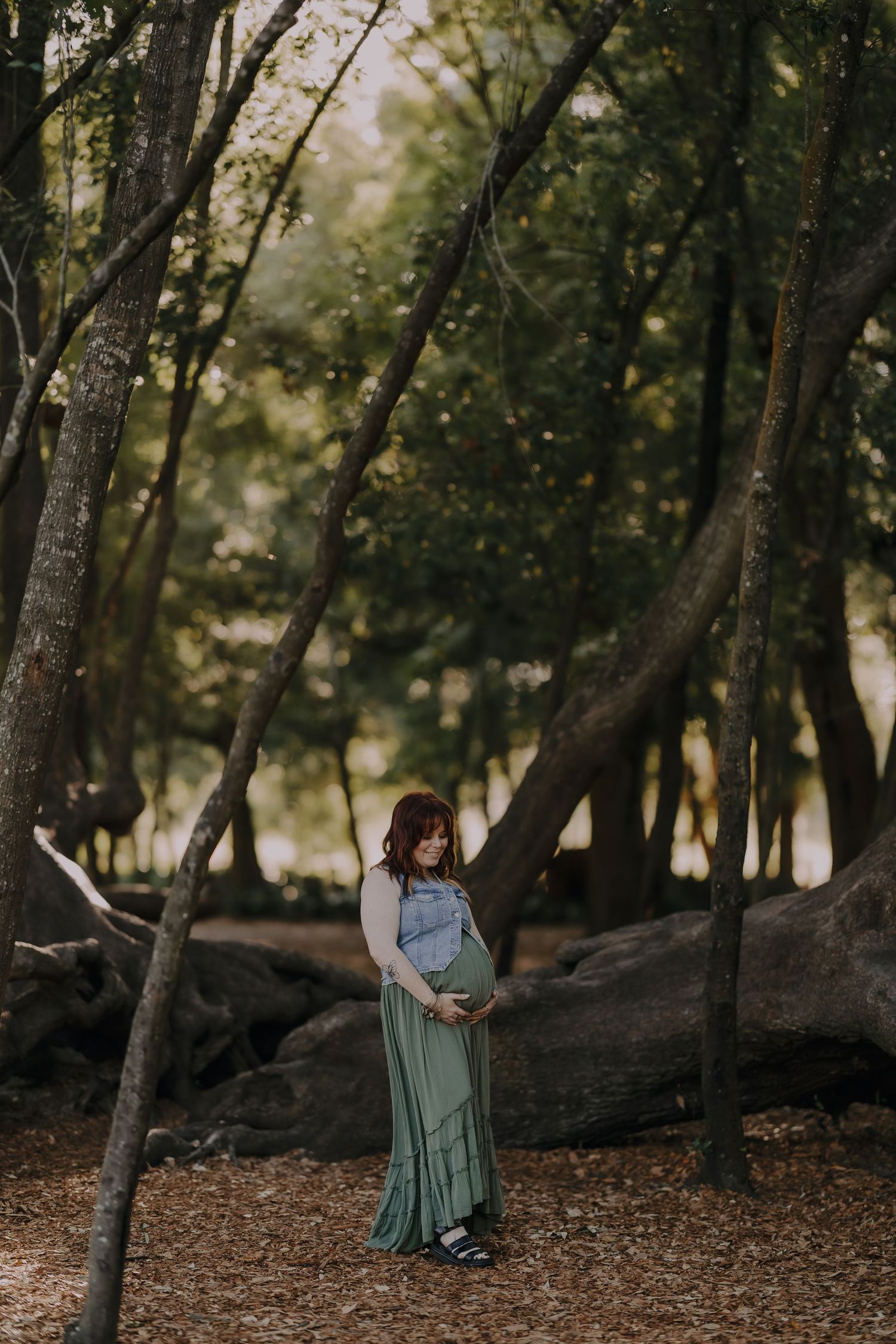 Pregnant woman in green maxi skirt and denim top cradling her belly among tall trees in a forest setting.