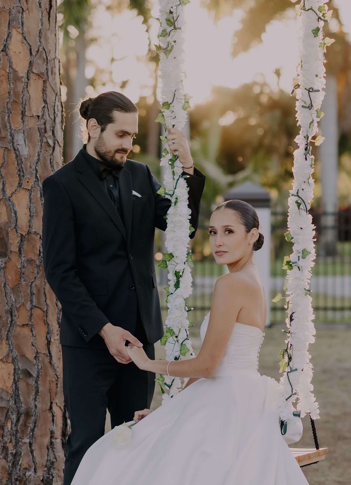 Bride in strapless white gown sits on floral swing while groom in black suit holds her hand beside a tree.