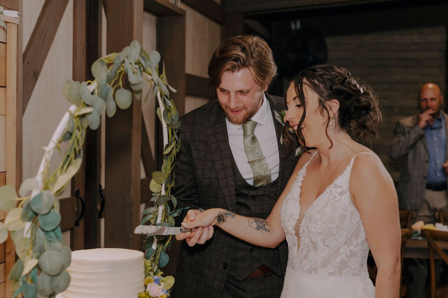Bride and groom cutting white wedding cake decorated with eucalyptus greenery at rustic indoor reception.