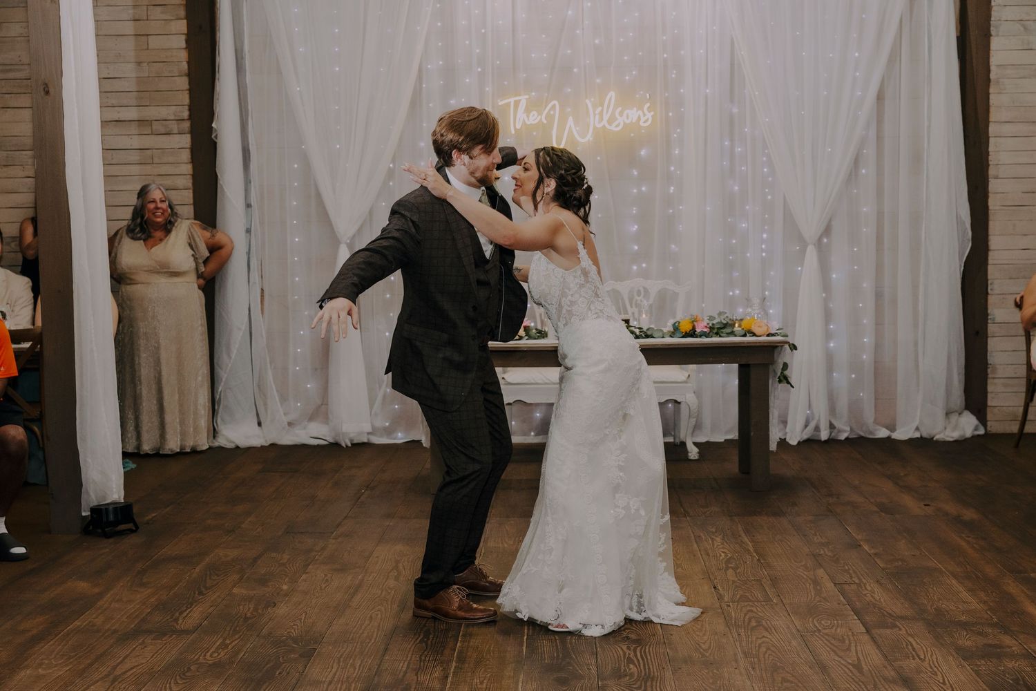 Bride and groom share their first dance at a rustic wedding reception with white drape backdrop and string lights.