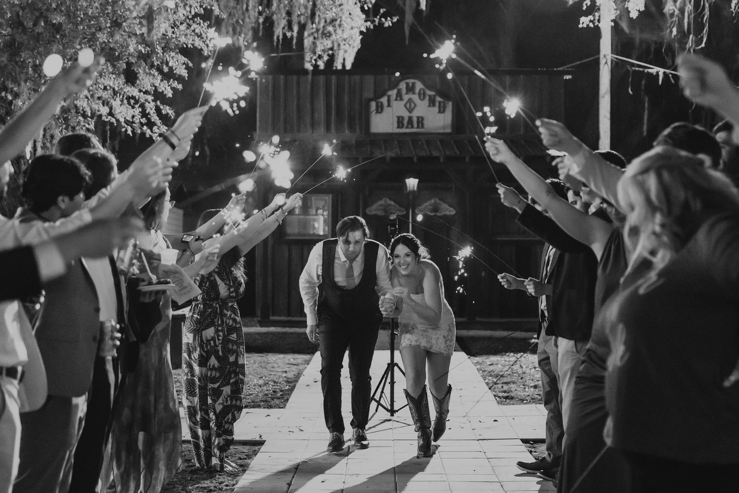 Bride and groom walk through sparkler tunnel surrounded by cheering guests at outdoor wedding reception.