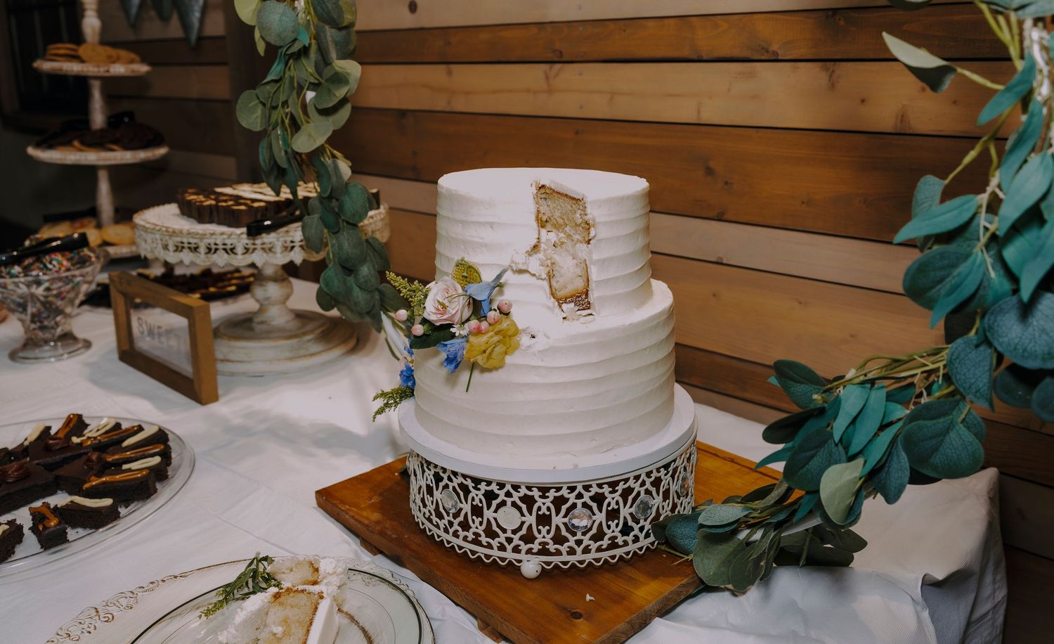 Two-tier white wedding cake with bear illustration and floral accents on decorative stand at rustic reception.