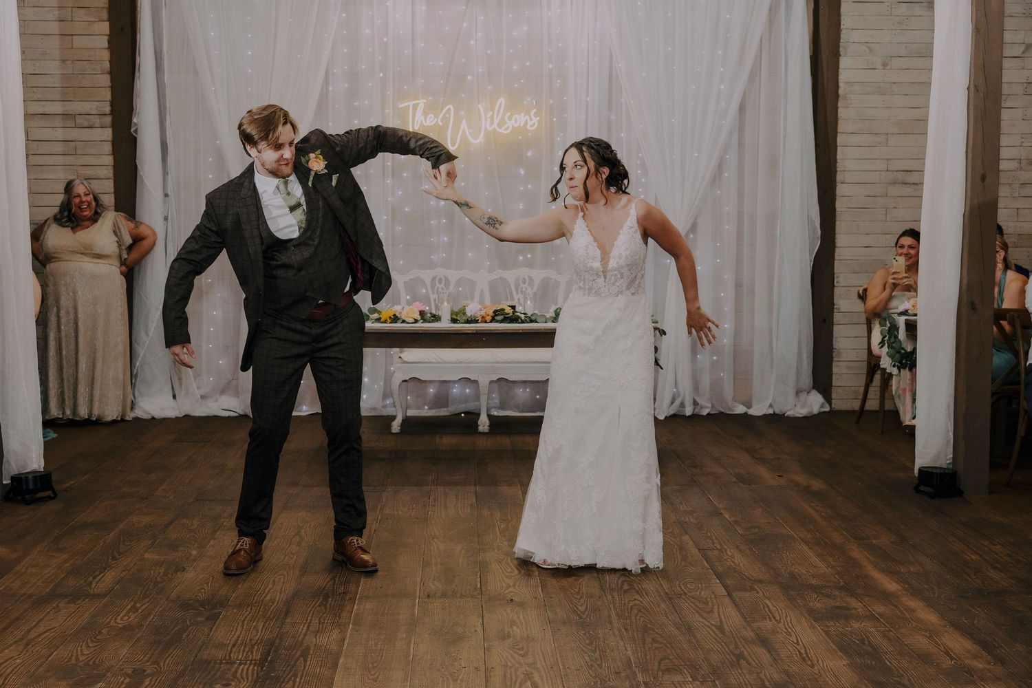 Bride and groom share a joyful first dance at rustic wedding reception with string lights backdrop.