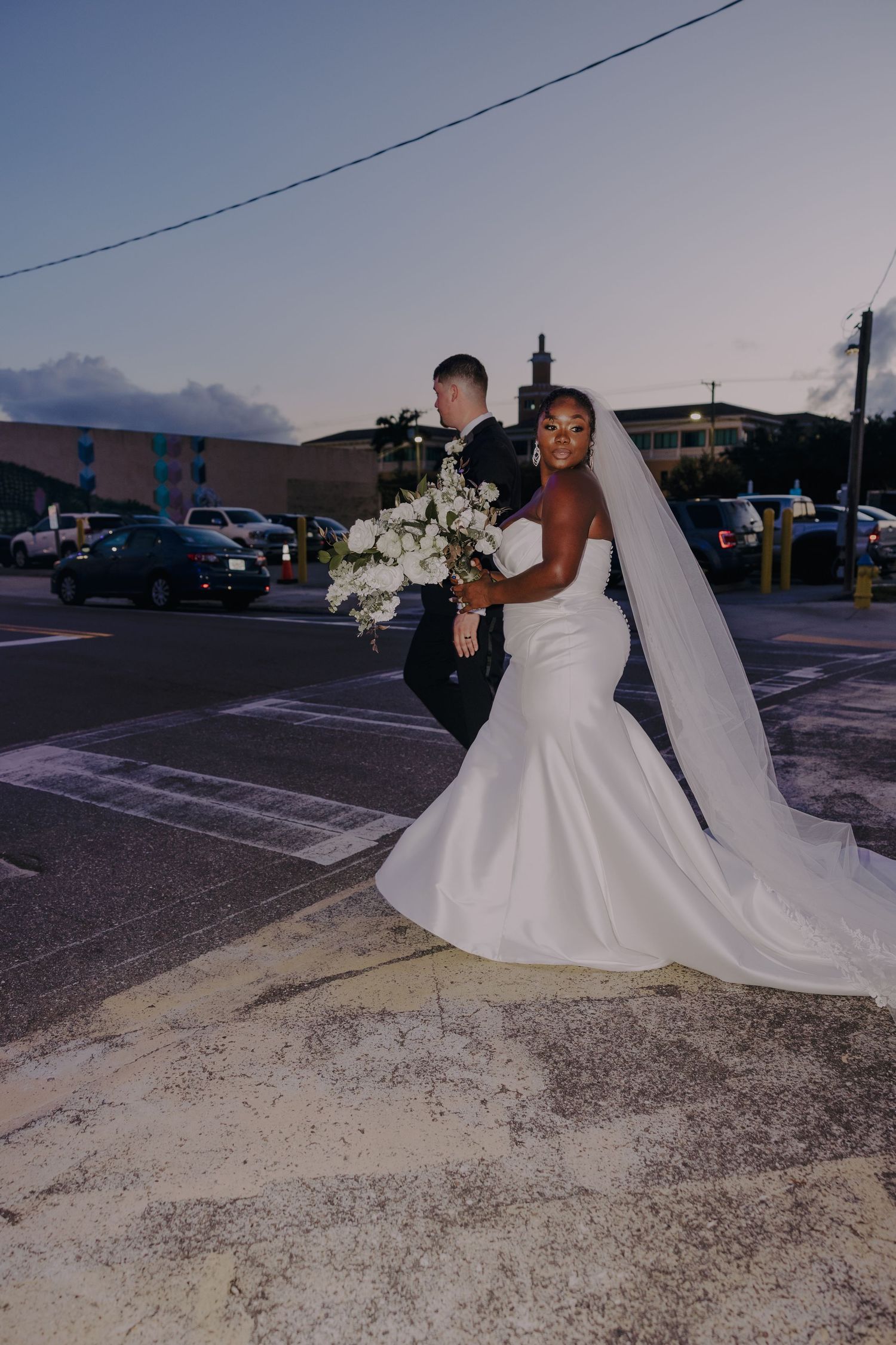 Bride in white mermaid gown and veil holds bouquet at dusk crosswalk, groom beside her.