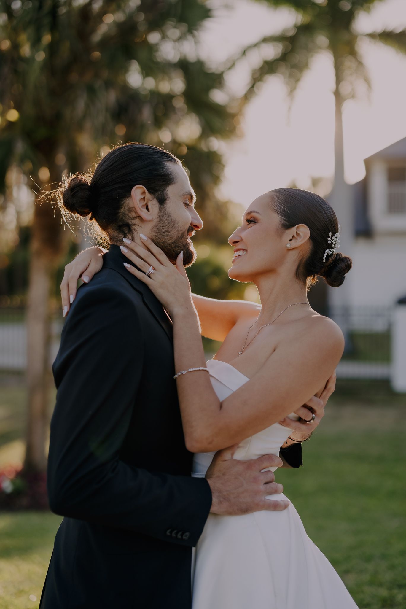 A bride and groom share a romantic embrace outdoors at golden hour, surrounded by palm trees at their wedding.