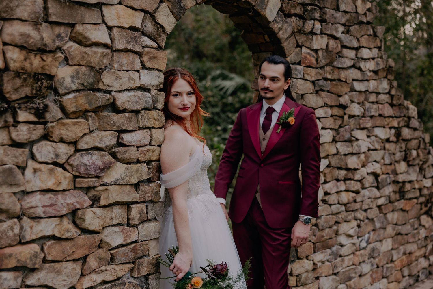 Bride in white off-shoulder gown and groom in burgundy suit pose by rustic stone arch wall outdoors.
