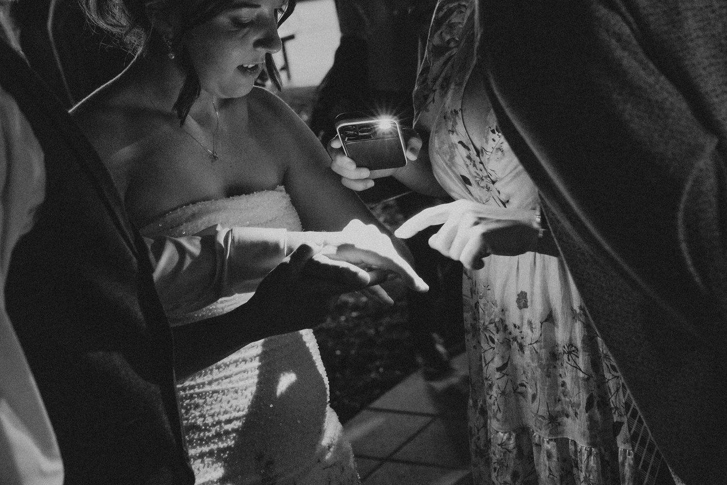 Black and white wedding photo of bride in strapless gown showing off ring to guests by phone flashlight.
