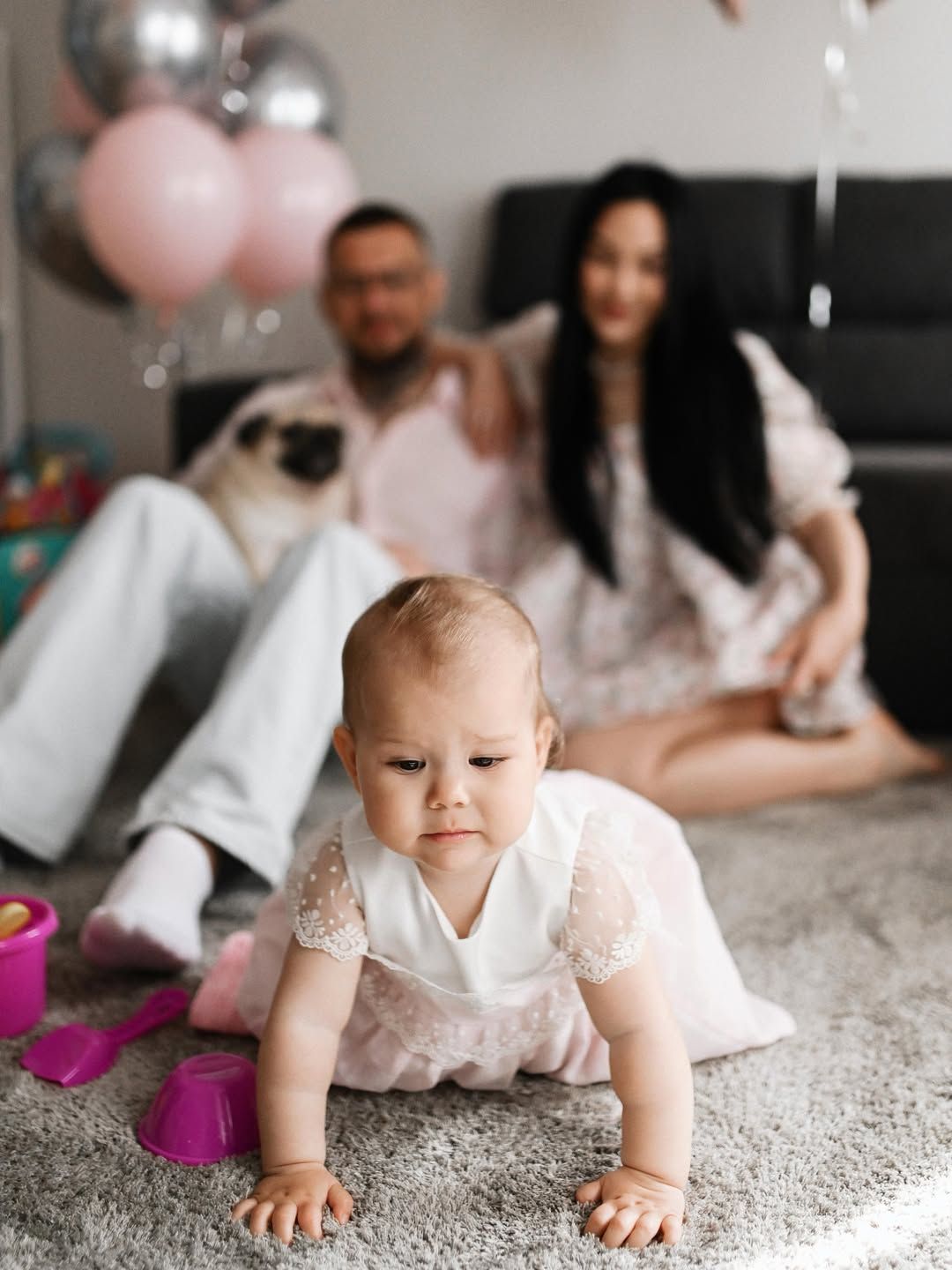 Close-up of a baby girl crawling on a gray carpet with parents and a pug dog sitting blurred in the background.