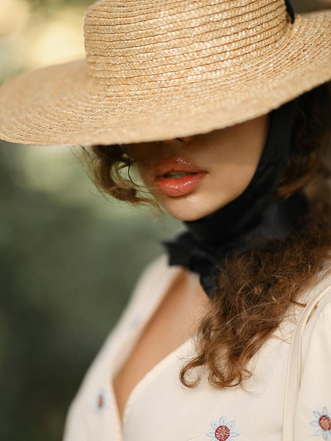 A close-up editorial portrait focusing on a woman's face and lips, shaded by the wide brim of a straw hat during a park photoshoot.