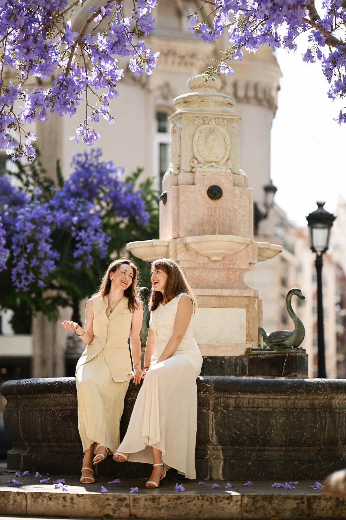 Two adult sisters laughing together by a stone fountain under blooming purple Jacaranda trees in Valencia.
