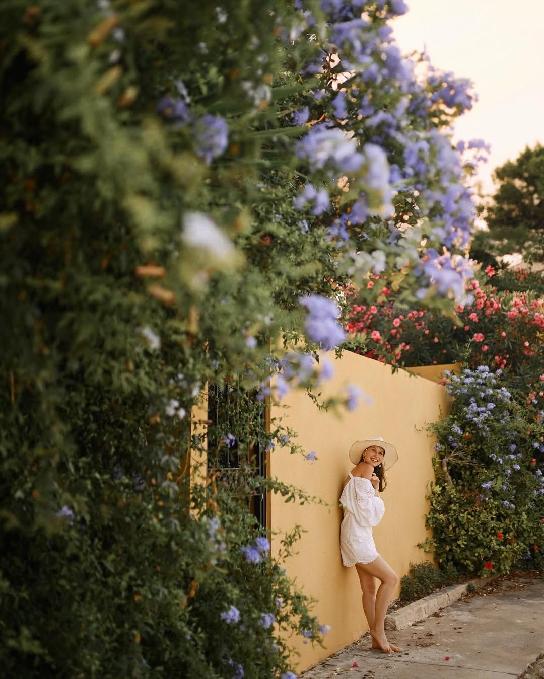 Smiling woman hugging herself against a bright mustard wall with blooming flowers and lush greenery in Port Saplaya village