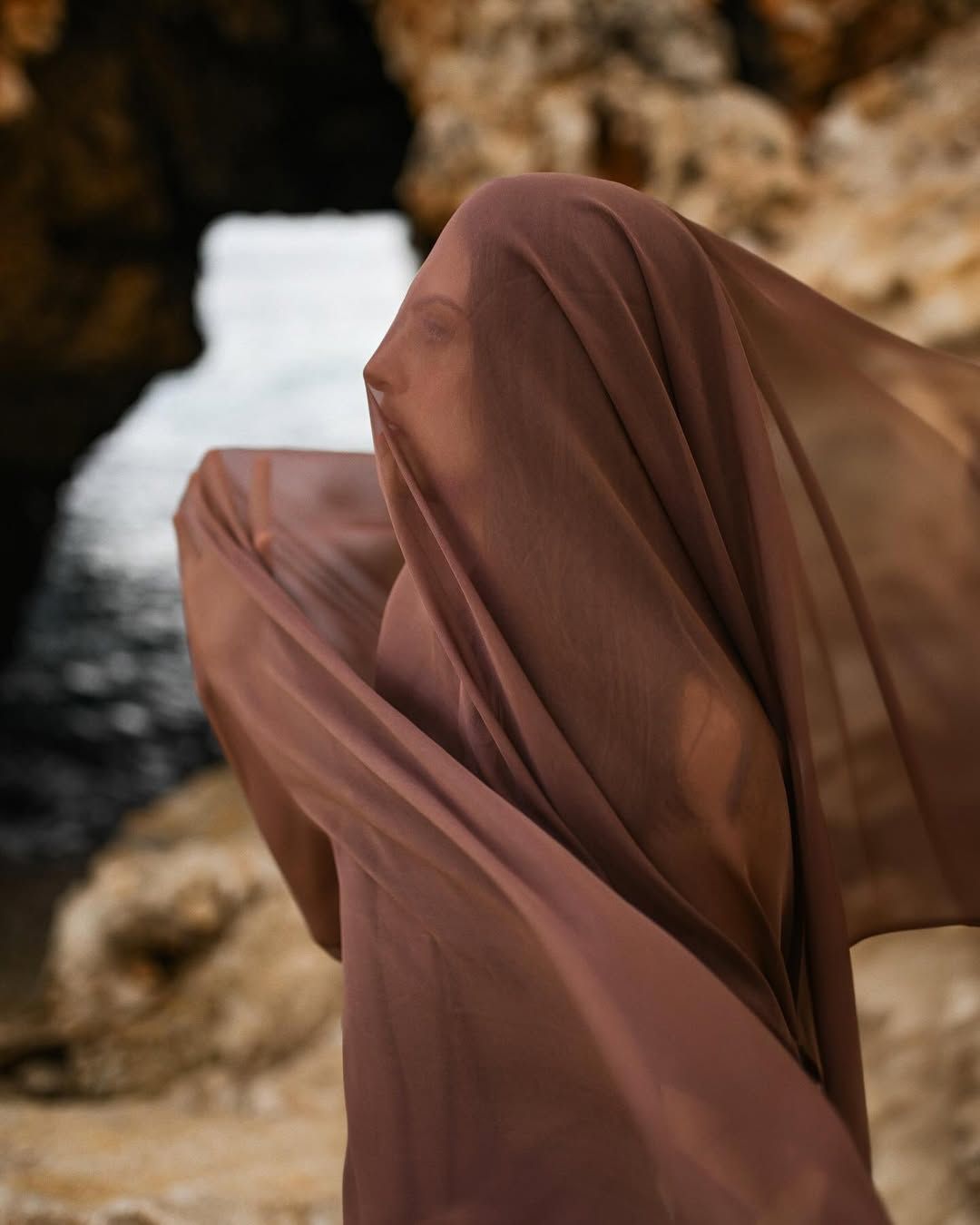 A cinematic portrait of a woman with a cloth draped over her face on the rocky shores of Cala Moraig beach.