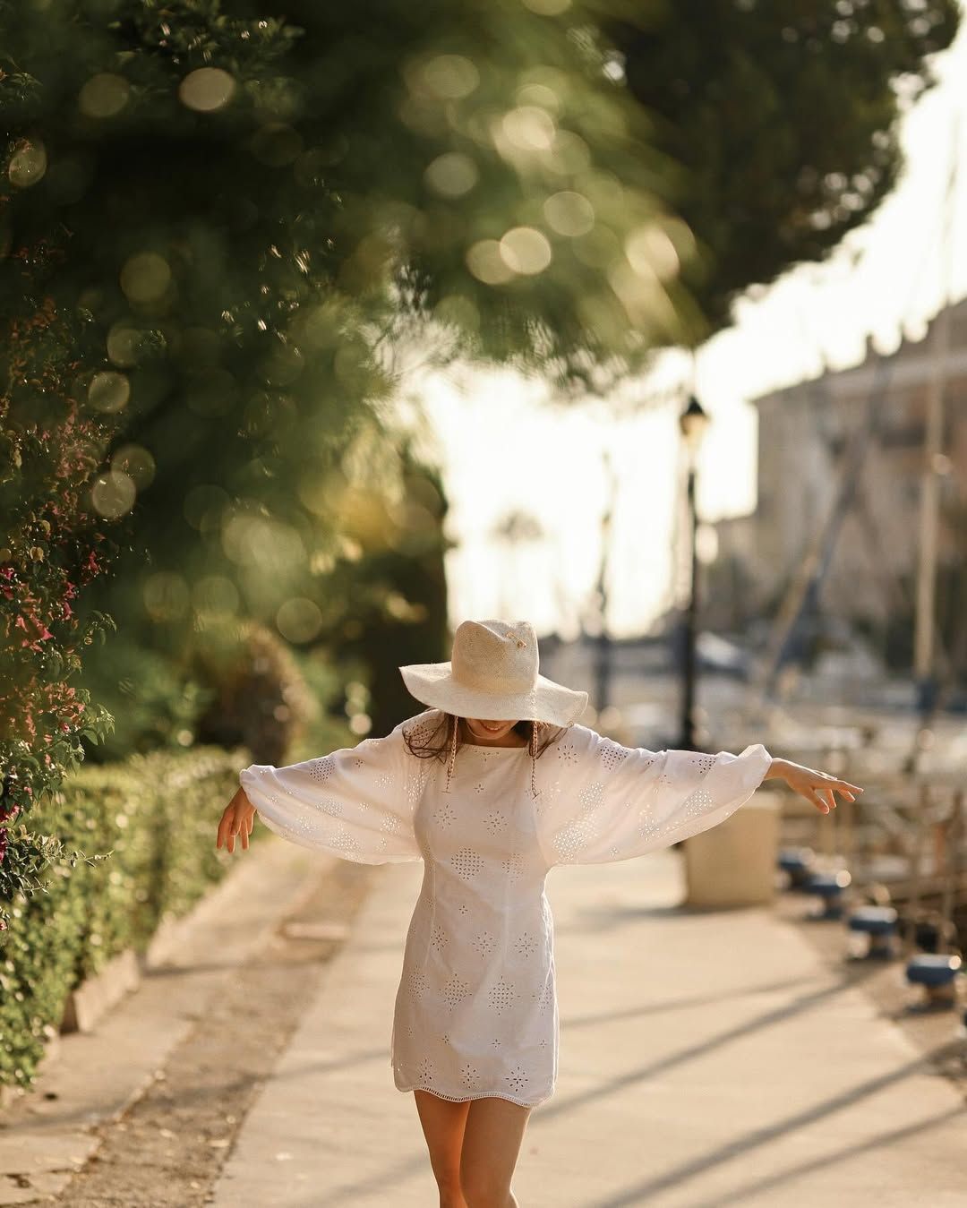 A relaxed woman stands with arms wide open, her face covered by a sun hat, against the scenic background of boats in Port Saplaya