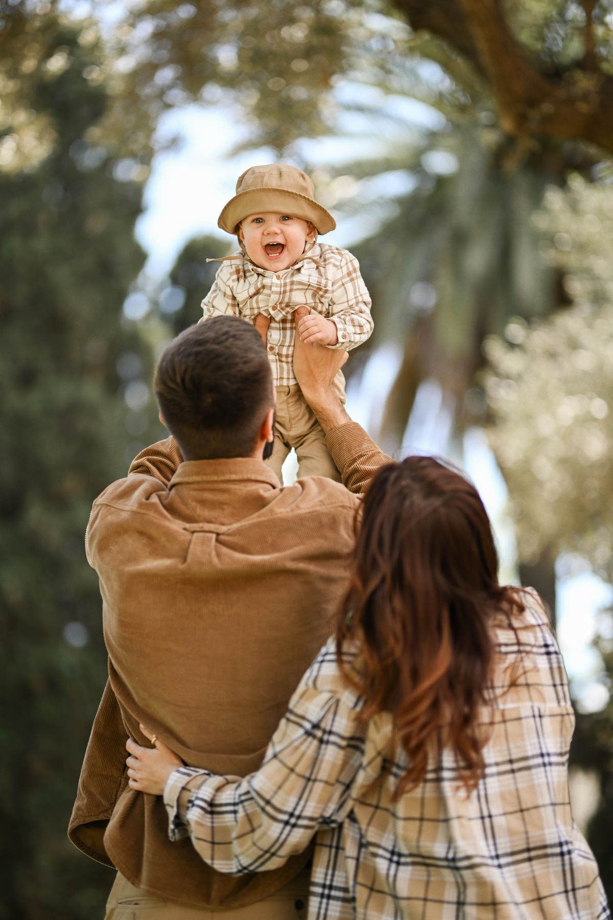 Father playfully lifting his laughing toddler son in the air during a family photoshoot in Viveros Park, Valencia.