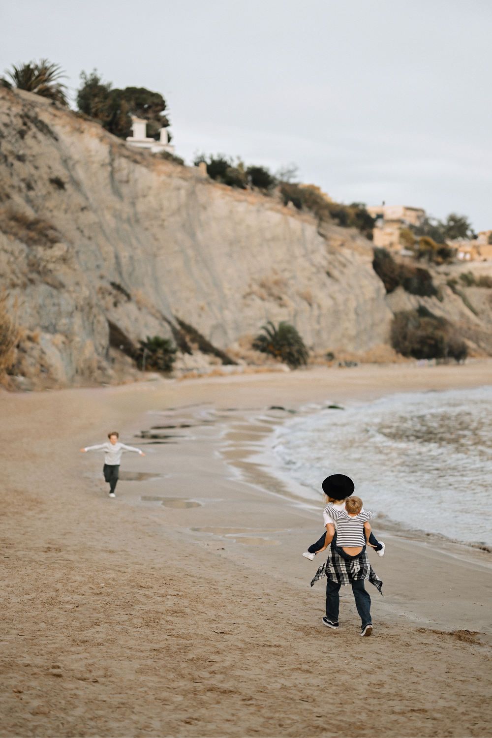 A mother giving her son a piggyback ride on the beach while her other child runs ahead during a lifestyle session in Calp