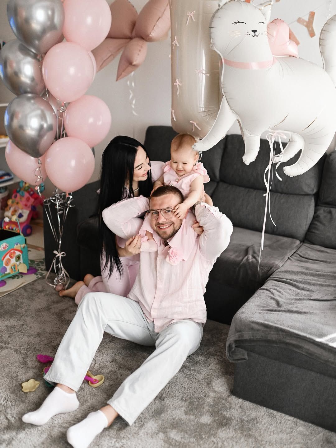 Happy father holding his baby daughter on his shoulders with mom nearby on the living room floor surrounded by balloons.