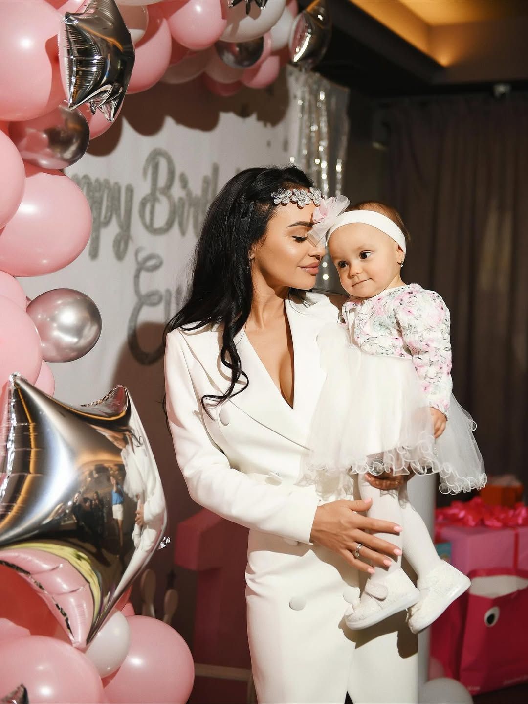 Mother holding her baby daughter at a bespoke birthday photo zone styled with pink and silver balloons.