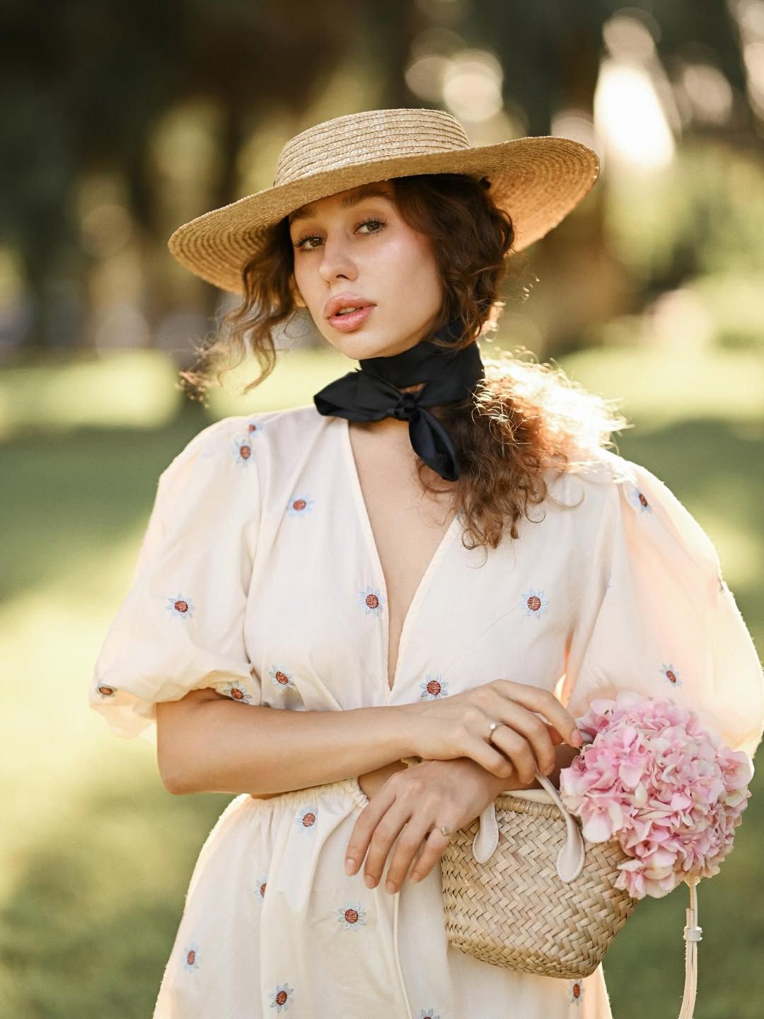 An elegant medium portrait of a woman holding a small basket with pink flowers, posing in the soft light of a beautiful park.