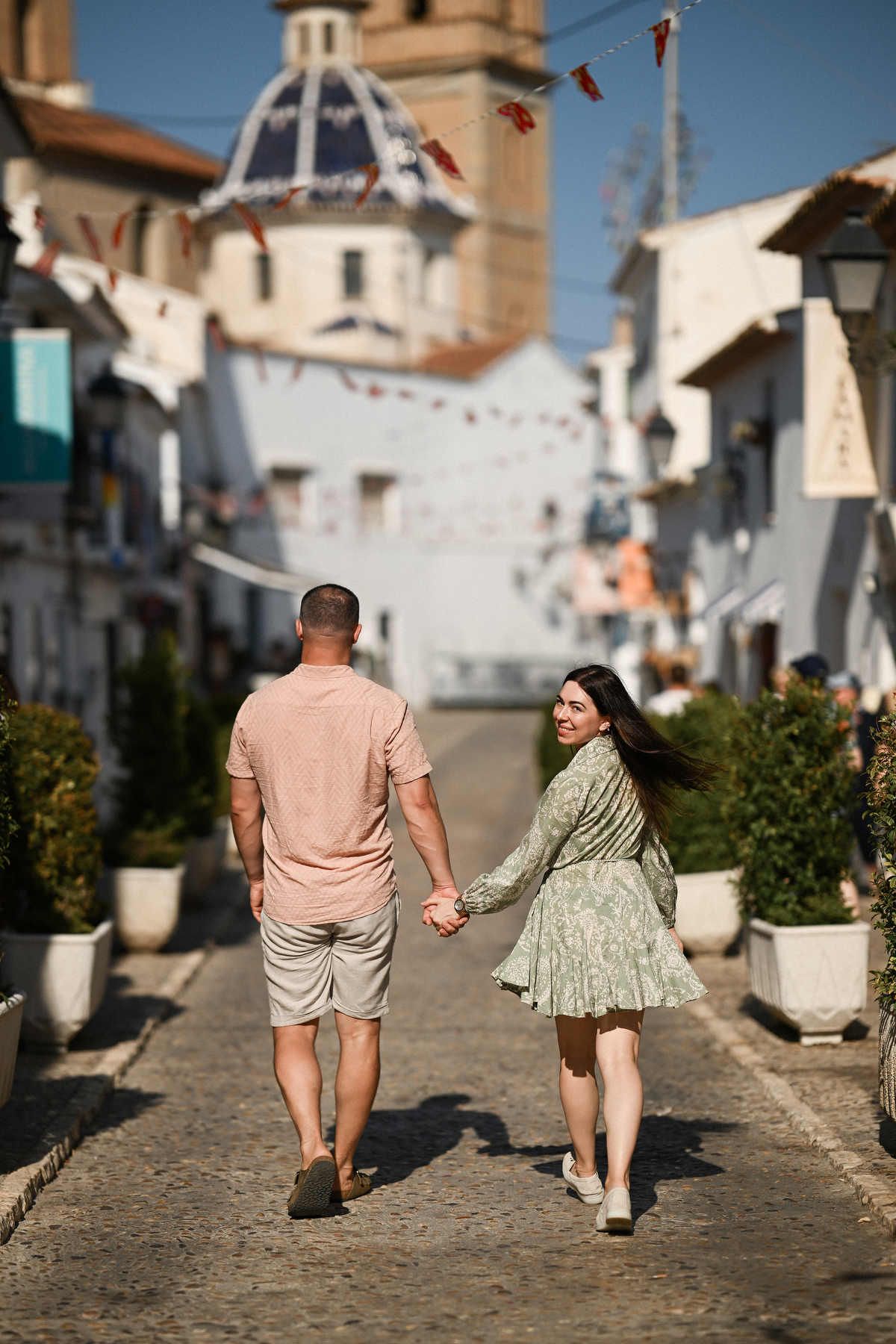 Parents holding hands walking towards the iconic blue domes of Altea; the mother looks back at the camera.
