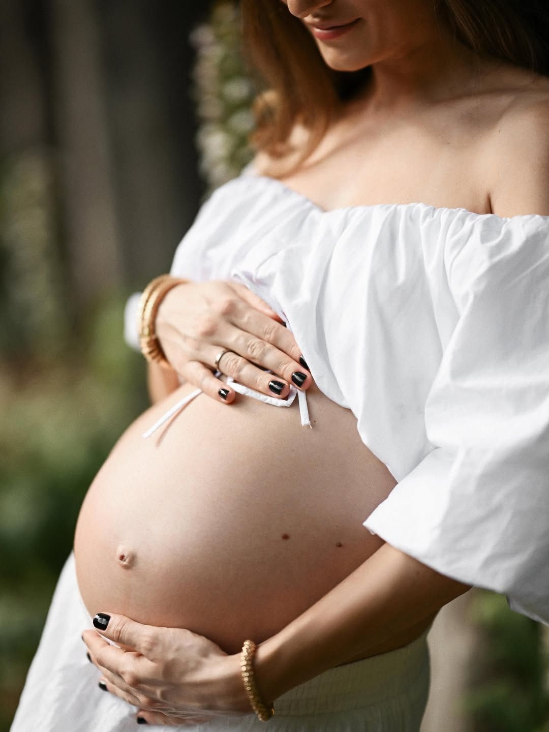 Close-up of an expecting mother's hands gently holding her pregnant belly in a white dress during a morning portrait session