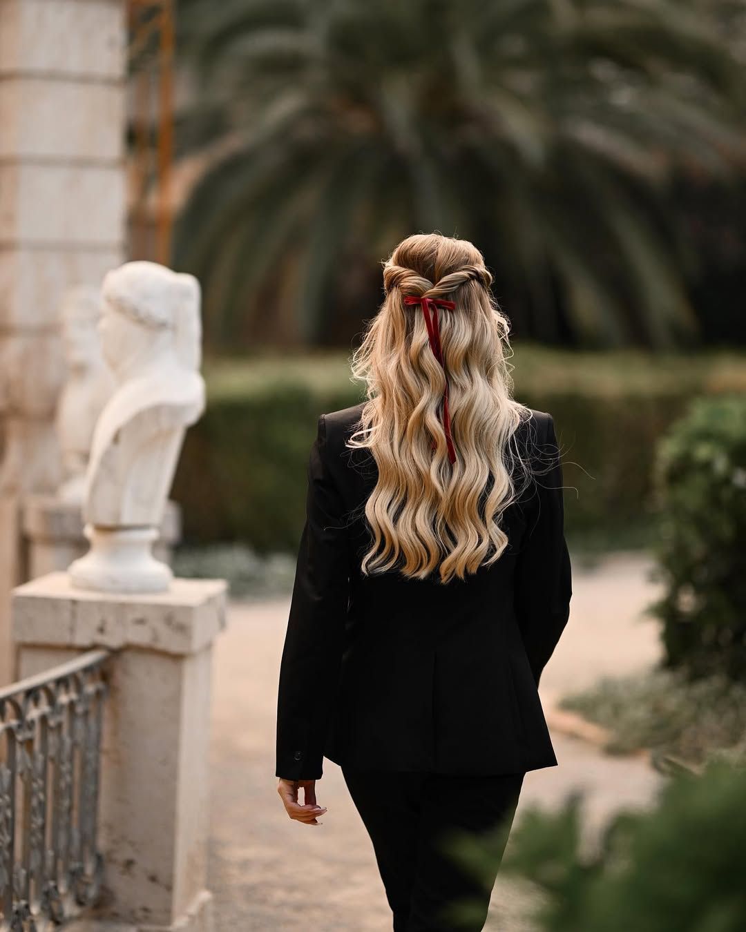 Back view of a blonde woman walking past classical marble statues in the geometric paths of Monforte Gardens.