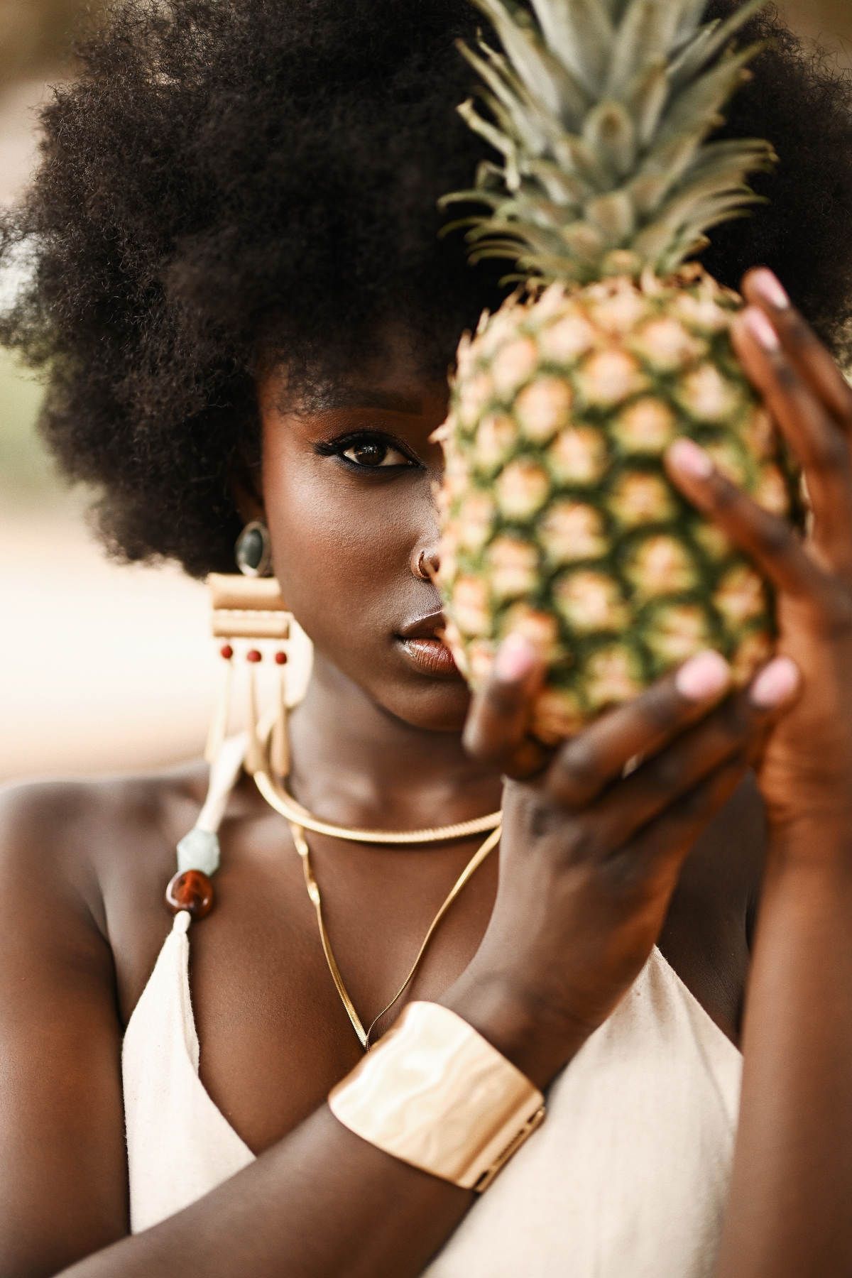 Striking close-up portrait of a model with an afro holding a pineapple, showcasing bold gold cuffs and ethnic styling in Valencia.