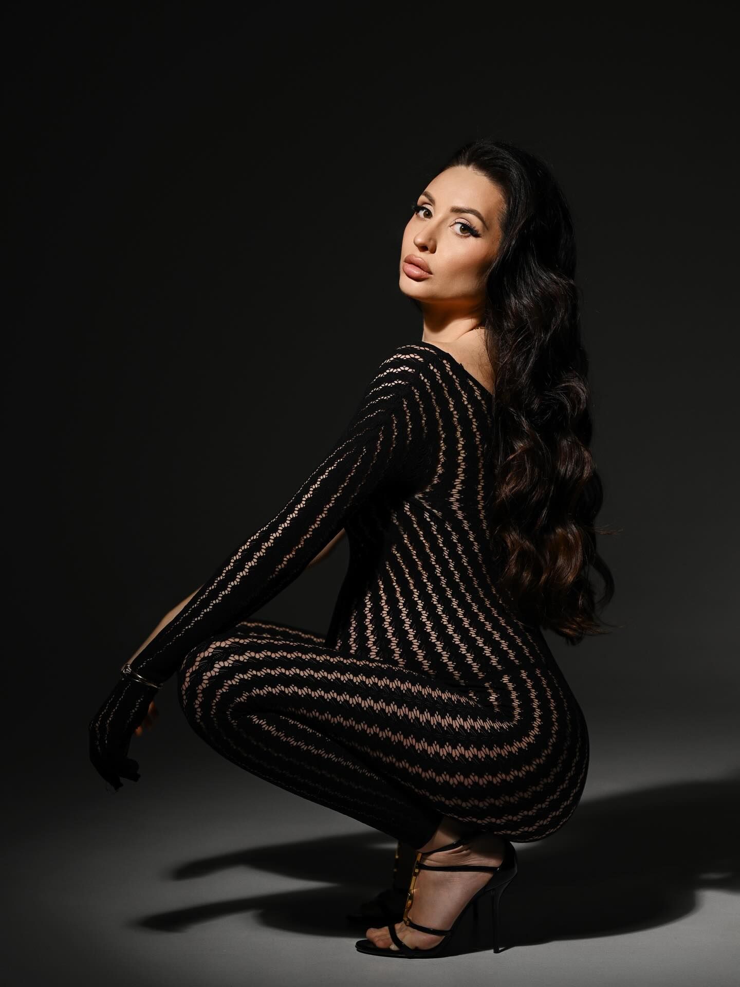 Elegant pregnant woman posing in a dimly lit studio wearing delicate sheer fabric.