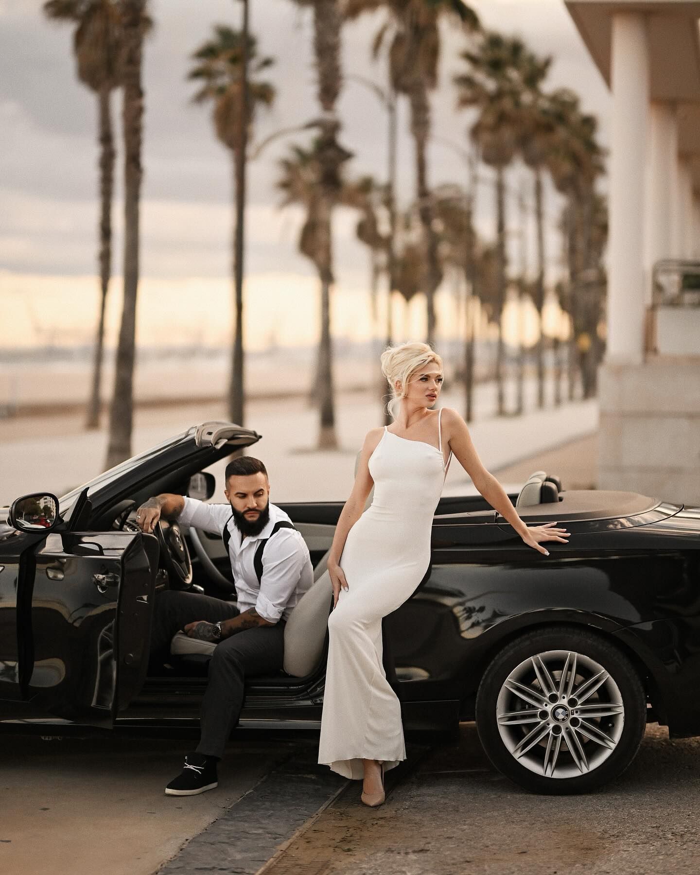 A bride in a sleek white dress posing next to a black convertible car with tall palm trees and a sandy beach background in Valencia.