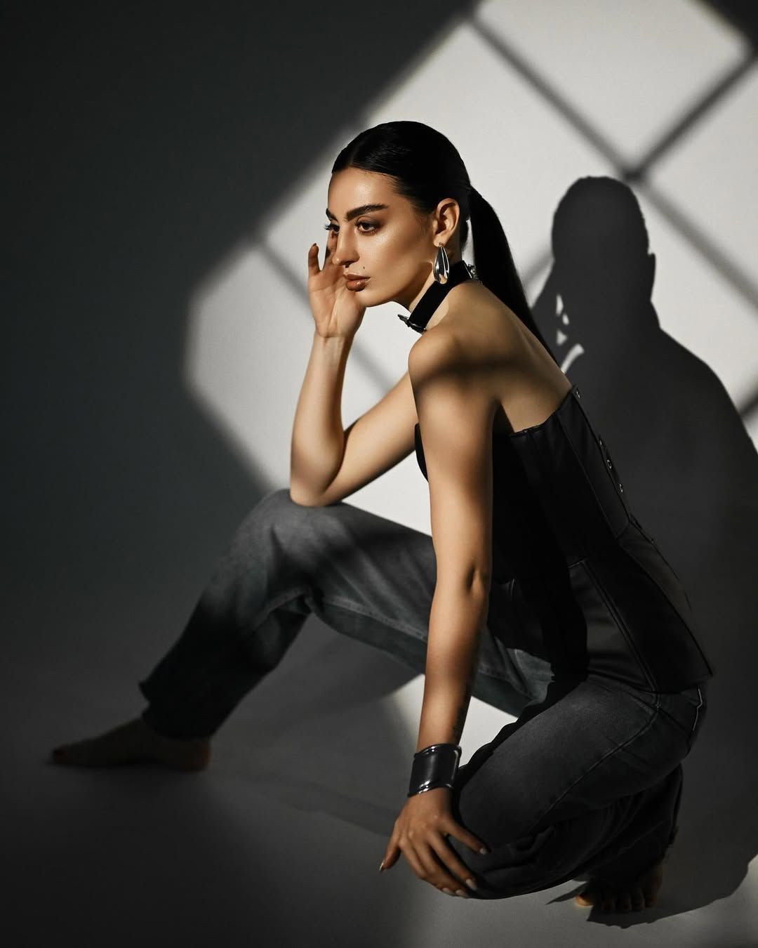 A side profile studio portrait of a woman posing against a wall with window shadow patterns, creating a minimalist fashion aesthetic.
