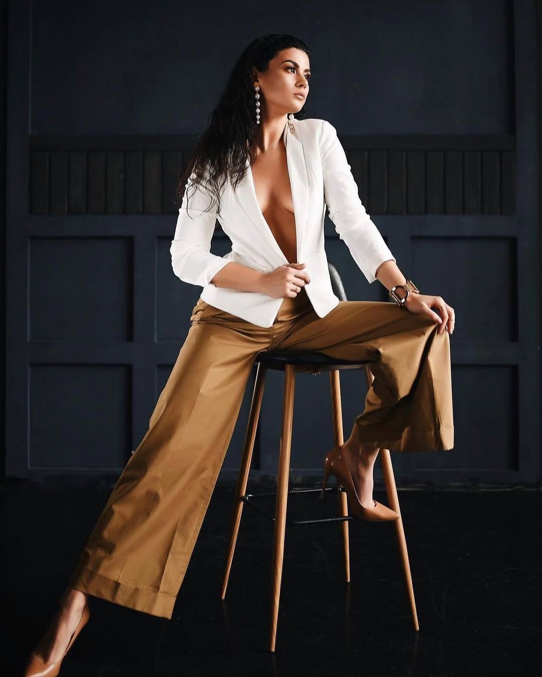 Editorial boudoir portrait in Valencia. A woman sits on a high stool wearing wide brown trousers and a white blazer styled elegantly bare.