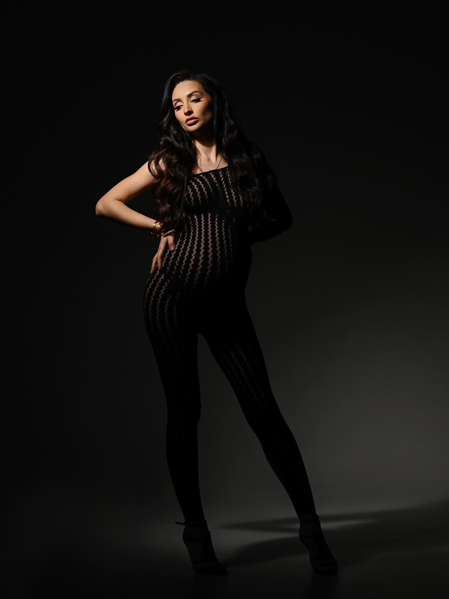 Pregnant woman in a sheer bodysuit standing in a minimalist studio with dramatic lighting.