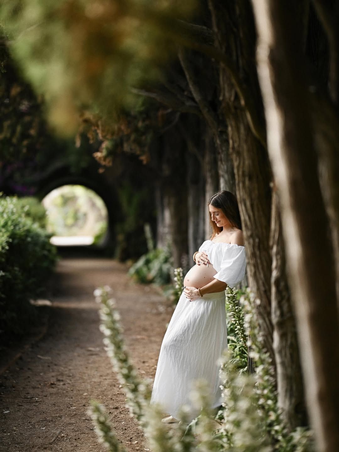 Pregnant woman in a white dress leaning against a tree along a shaded ivy-covered path in Jardines de Monforte, Valencia