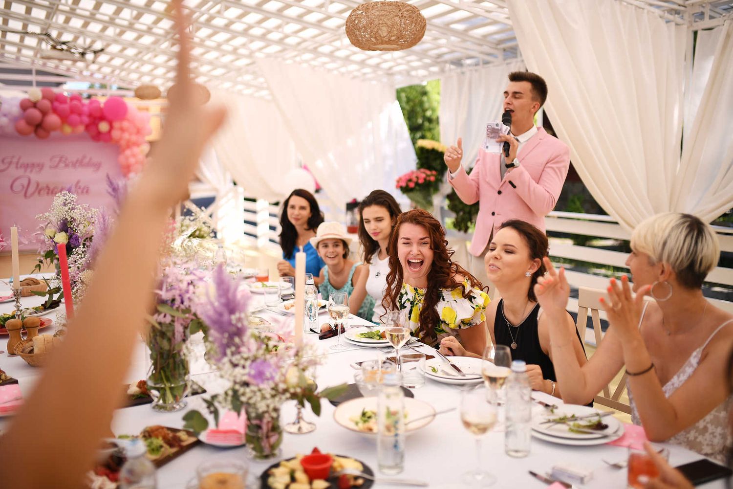 Guests laughing at a beautifully decorated event table while an entertainer in a pink blazer hosts the party show program.