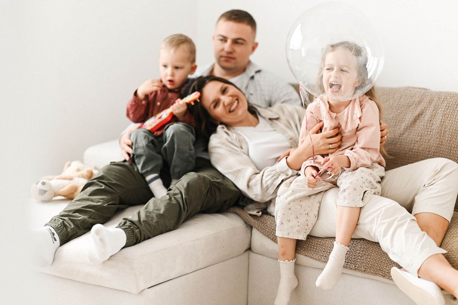 Parents sitting on a sofa with their two children, who are playing with a clear balloon and a toy, during a fun home photography session.