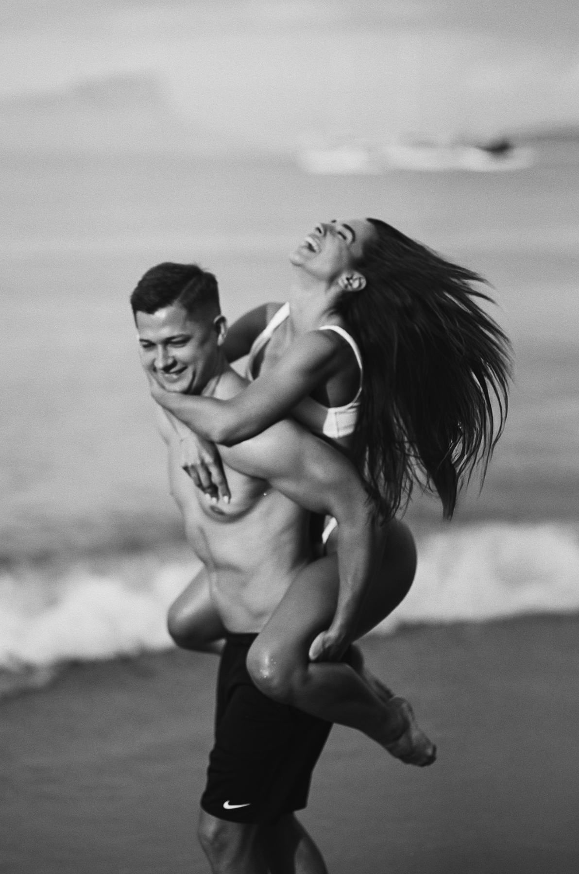 Candid black and white portrait of a man giving a woman a playful piggyback ride on a beach in Moraira