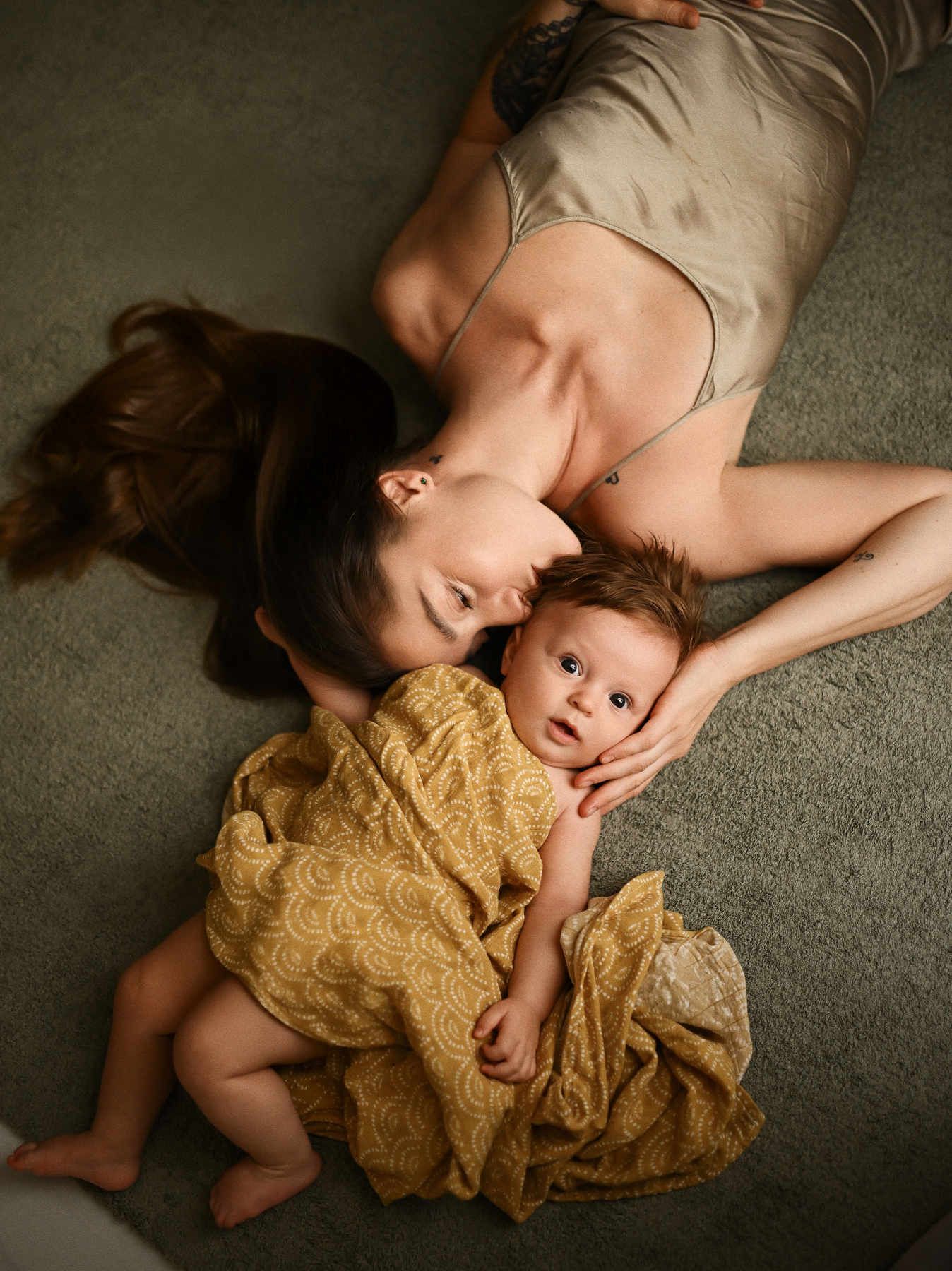 A soft, lifestyle photograph of a mother kissing her baby's head while resting together in a cozy home environment.